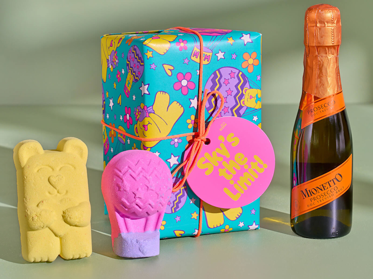 Ein bunt verpacktes Geschenkset von LUSH mit zwei farbenfrohen Badebomben in Form eines knallig gelben Kuschelbären und eines pinken Heißluftballons. Daneben steht ein Piccolo Prosecco.