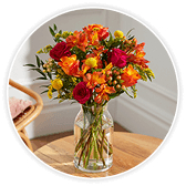 A glass vase of vibrant red, orange, and yellow flowers including roses and freesia, arranged with greenery, standing on a wooden table.