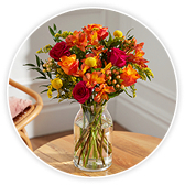 A glass vase of vibrant red, orange, and yellow flowers including roses and freesia, arranged with greenery, standing on a wooden table.