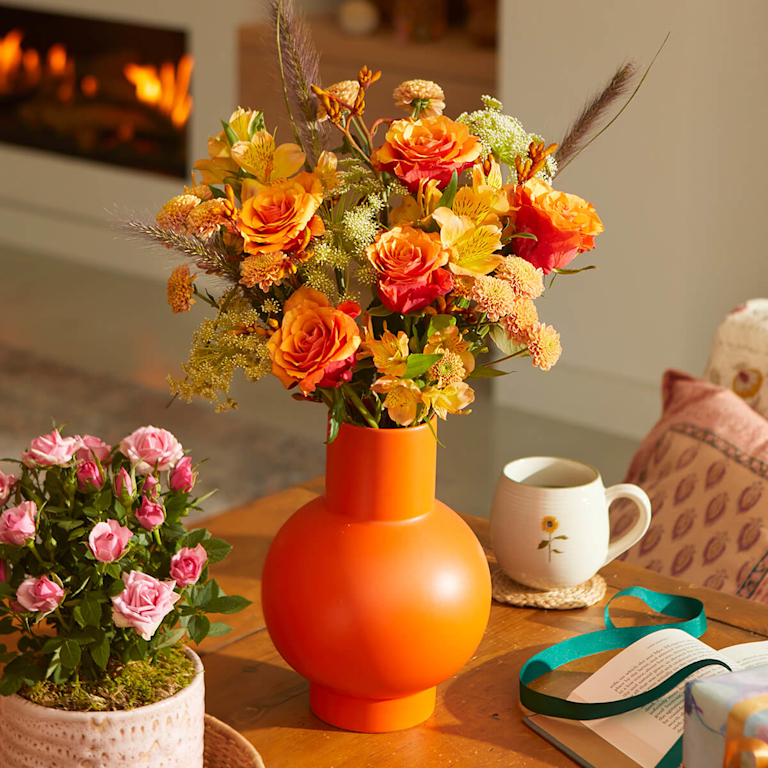 A beautiful autumnal bouquet in an orange vase, filled with orange roses and yellow alstroemeria/ On the right, a mug sits next to an open book with a green ribbon.