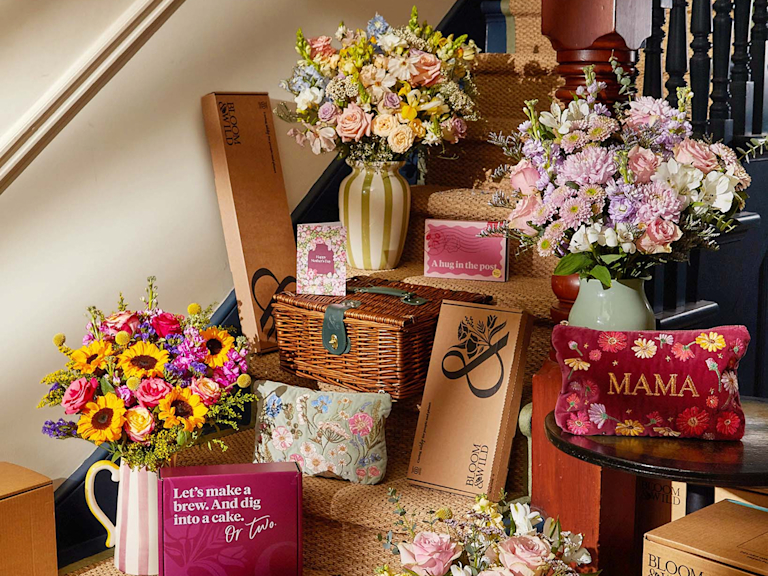 A staircase styled with vibrant Mother’s Day gifts. Features sun-drenched sunflowers, pastel roses, and wild meadow bouquets in striped vases alongside a wicker hamper, velvet ‘MAMA’ embroidered pouch, and Bloom & Wild gift boxes.