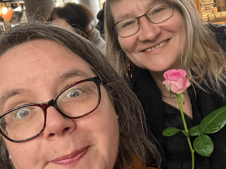 Katherine and Judy, two women wearing glasses, smile at the camera in a cafe. Katherine holds a pink rose.
