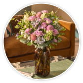 Eine braun gemusterte Glasvase ist mit üppigen Rosen, Dahlien und Löwenmäulchen in Rosa-, Flieder- und Cremetönen gefüllt. Der Strauß steht auf einem Tisch vor einem braunen Sofa in warmem Tageslicht.