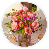 Eine pink getönte Glasvase mit einem üppigen Bouquet aus rosafarbenen Rosen, Nelken und Gladiolen steht auf einem gedeckten Tisch.