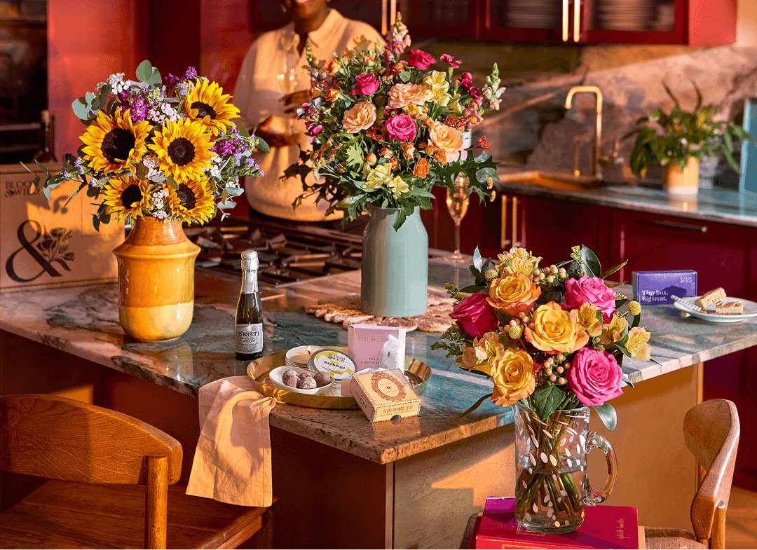 Three bouquets on a marble kitchen island: sunflowers in a yellow vase, pink and peach roses in a sage vase, and pink and yellow roses in a glass jug, with prosecco, chocolates and gifts in warm light.