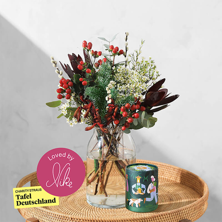 Ein rustikaler Blumenstrauß mit Beeren und Tannenzweigen steht in einer Vase auf dem Beistelltisch. Daneben ist eine runde, festlich illustrierte Schachtel mit Lebkuchen zu sehen.
