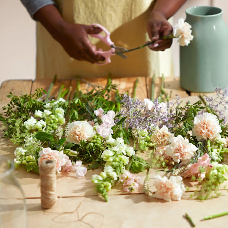 A person arranging a bouquet of pink roses, purple statice, pink carnations, and yellow tulips on a table.