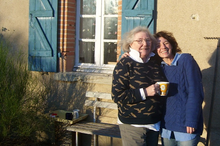 Sarah puts her arm around her mum Ann, smiling and standing outside a country cottage in summer.