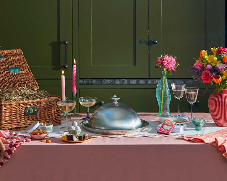 An animated dinner setting featuring a wicker hamper, champagne, and a vibrant pink bouquet. A silver cloche is lifted to reveal treats, creating a sense of surprise and celebration with a warm, floral-scented ambiance.