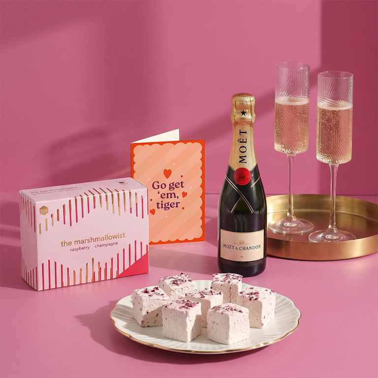 Celebration gift set with Moët Champagne, flutes, powdery pink raspberry champagne marshmallows, and a card reading "Go get 'em, tiger" on a bright pink backdrop.