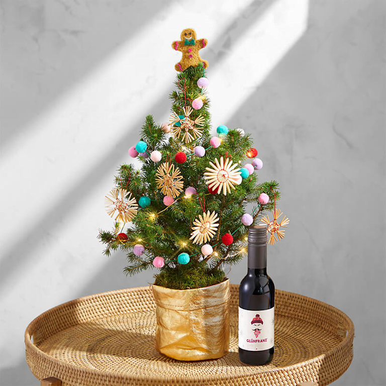 Ein kleines Weihnachtsbäumchen geschmückt mit filigranen Strohsternen und einer bunten Pom-Pom-Girlande steht in einem goldenen Übertopf auf dem Tisch. Daneben ist eine Piccolo-Flasche mit rotem Glühwein von Schorlefranz zu sehen.