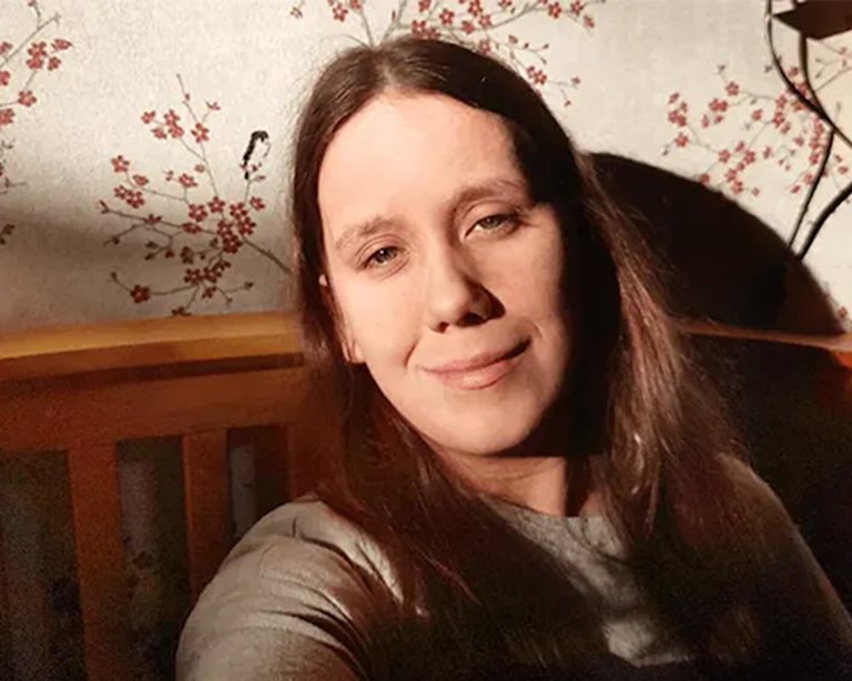 A young woman with long brown hair smiles as she rests against a wooden bed headboard. A slice of sun gently illuminates her face. 