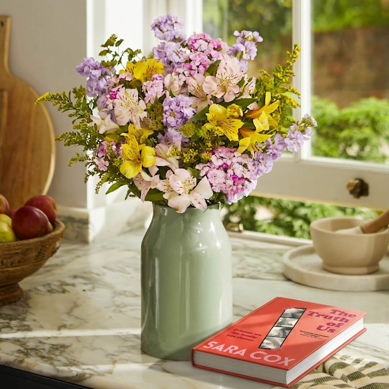 A bright kitchen countertop scene featuring a large, vibrant bouquet of purple, yellow, and pink flowers in a sage green ceramic vase. Lying flat on the marble countertop to the right of the vase is a hardcover book titled "The Truth of Us" by Sara Cox.