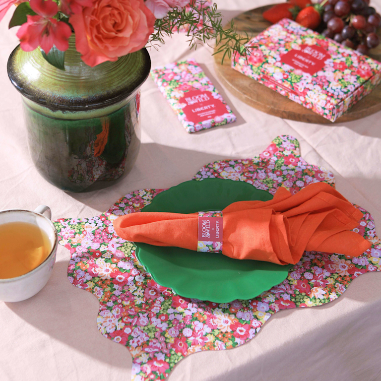 A beautifully set table featuring a small bouquet of pink roses in a green vase, a floral-patterned place setting with a bright orange napkin, a cup of tea, and a box of teabags with a matching floral design.