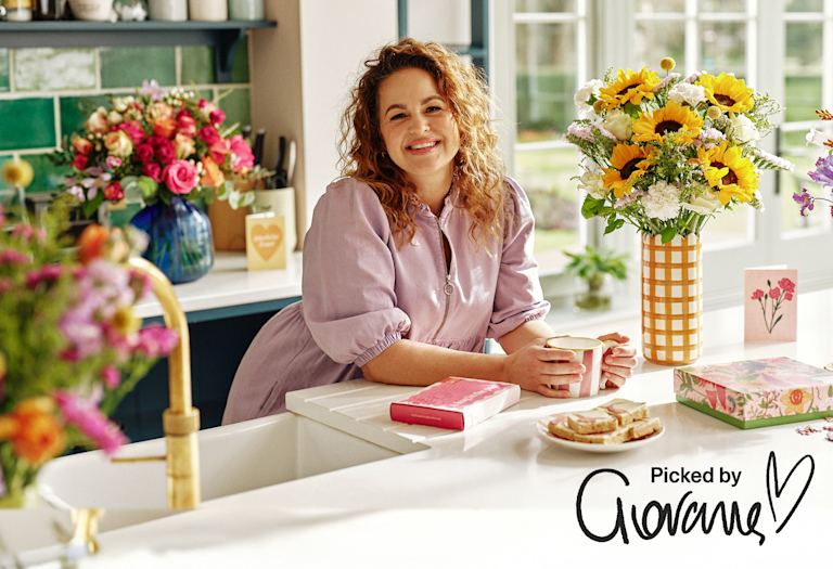 Smiling Giovanna Fletcher with bright sunflower and mixed flower bouquets and gifts on a sunny kitchen counter.