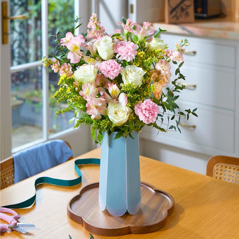 A lush bouquet of ruffled pink carnations, ivory roses, and delicate alstroemeria in a pale blue scalloped vase. Fresh eucalyptus and golden solidago add a wildflower feel. Displayed on a wooden tray with floristry scissors nearby.