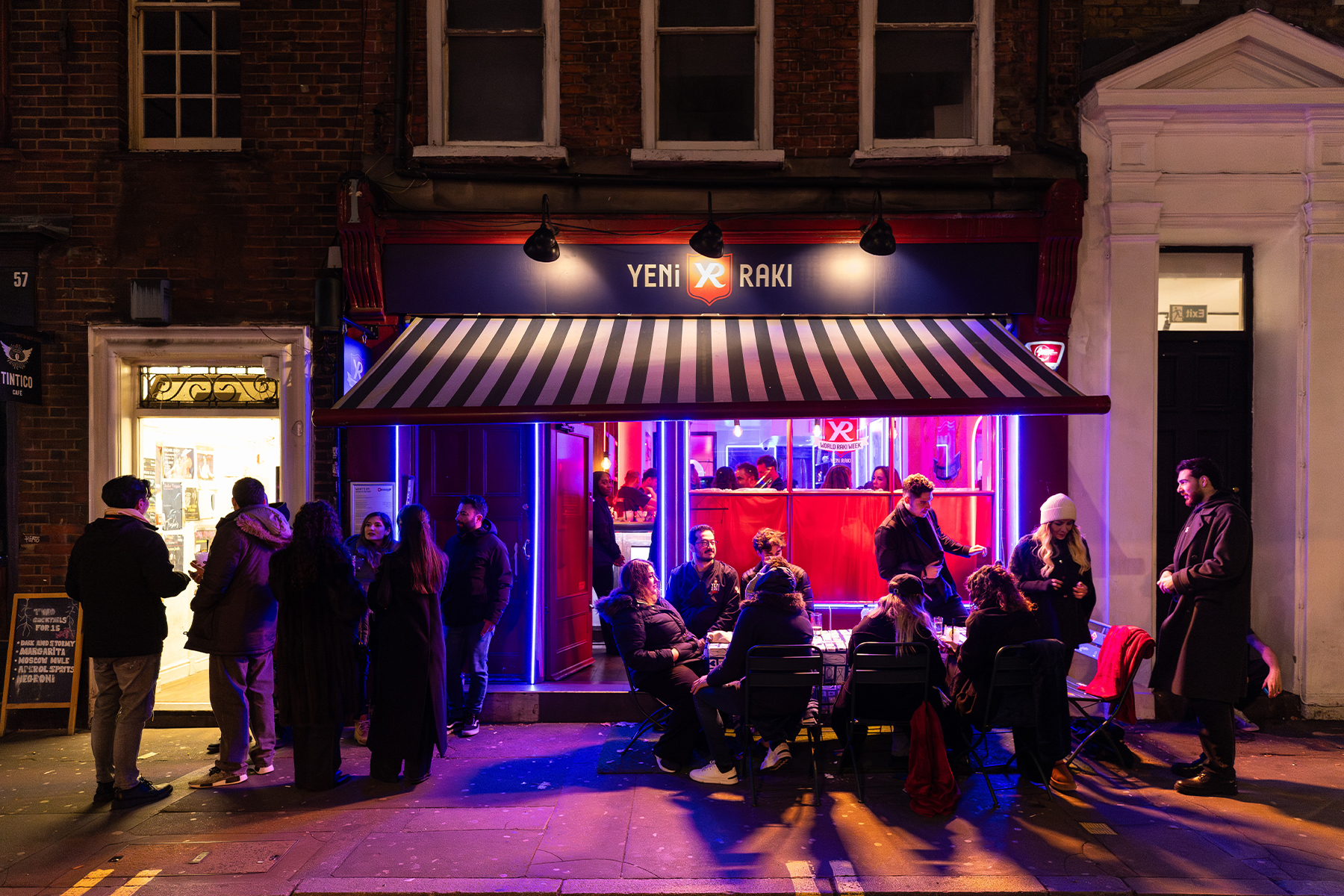 Nighttime street scene outside a lively Yeni Raki bar, featuring a striped awning and illuminated signage.