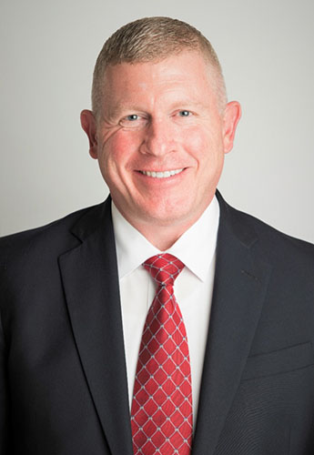 Older white man with short blonde hair wearing a dark suit with a white dress shirt and red tie.