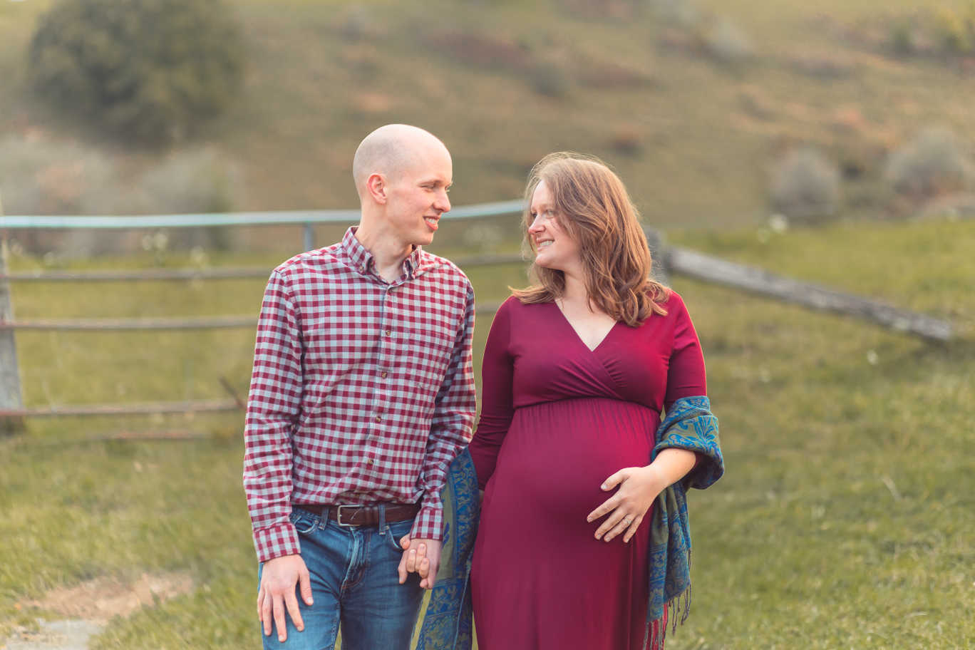 A man and woman holding hands looking at each other with a fence in the background. The woman is holding her pregnant belly.