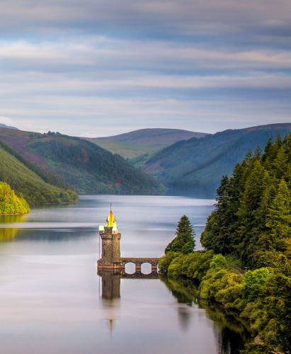 Sunset sunrise golden hour over Lake Vyrnwy reservoir surrounded green trees forest reflecting water tower panorama of welsh countryside