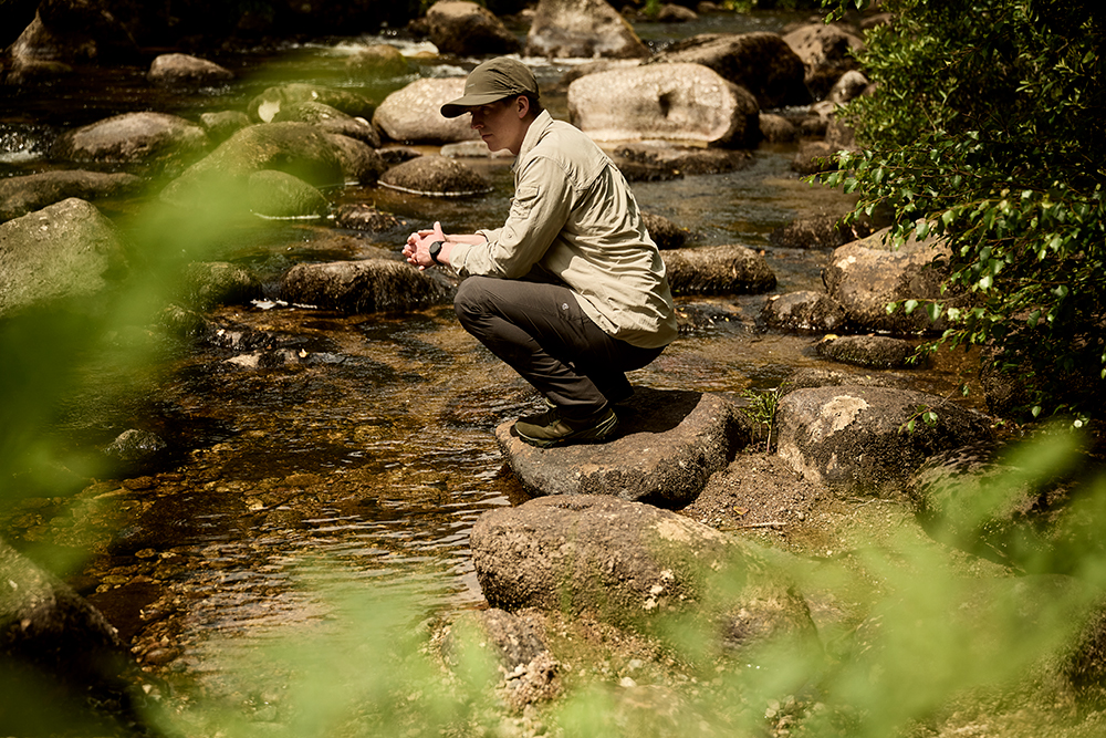 Man perched on a rock by a river wearing nosilife hat and jacket