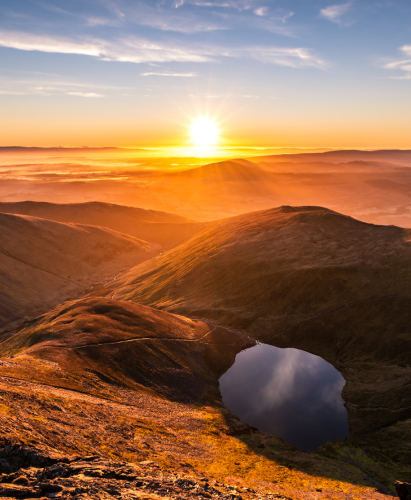 Stunning orange yellow sunrise from Sharp Edge on Blencathra, Lake District, Cumbria in Spring Summer with reflection in lake tarn and cloud inversion over mountains in the distance