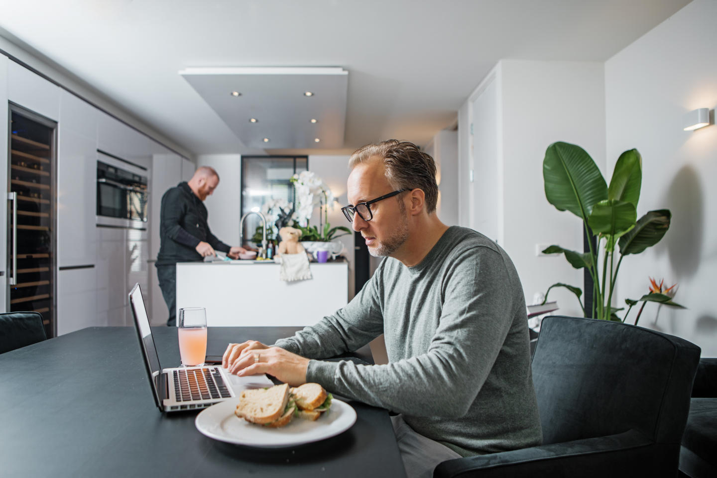 Man working on laptop on the kitchen table. Next to him is a sandwich on a plate, and we can see into the kitchen, where there's a man doing dishes