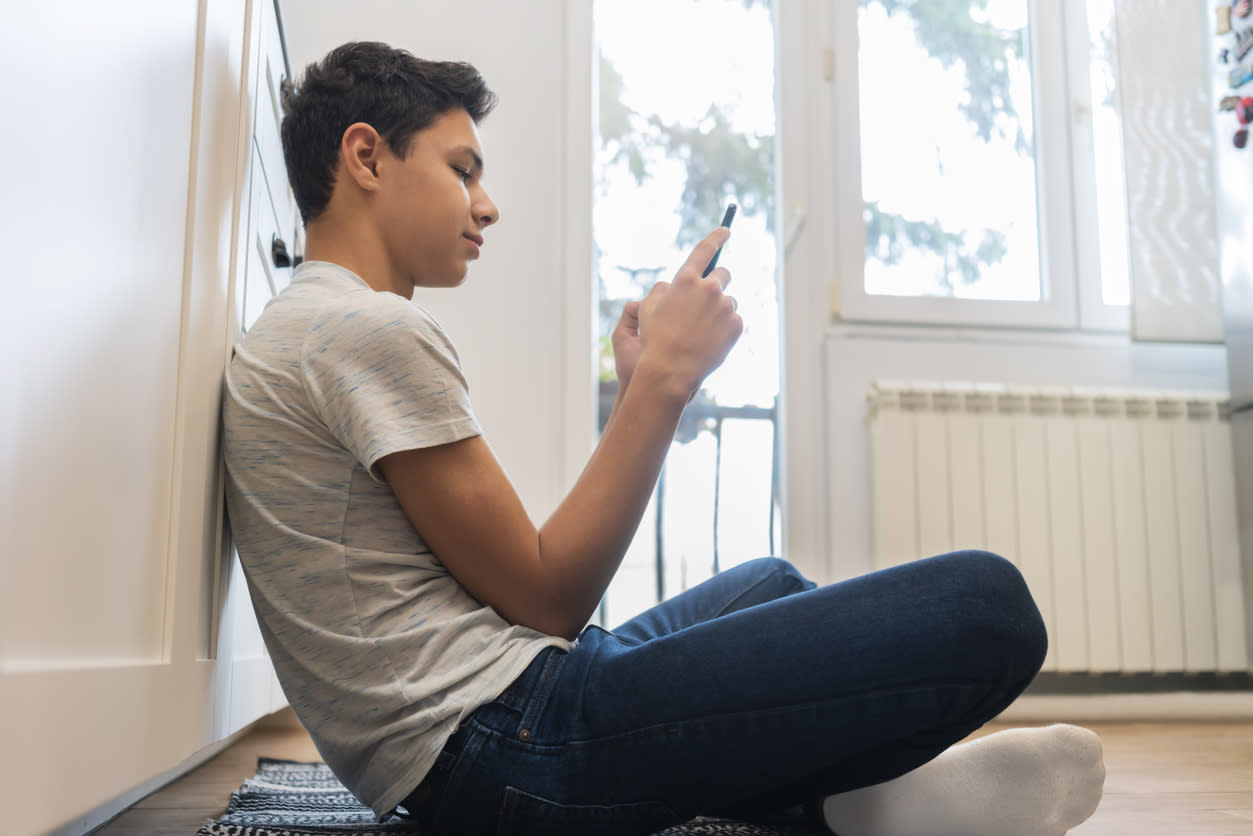 A teen boy sits on the floor looking at his smartphone