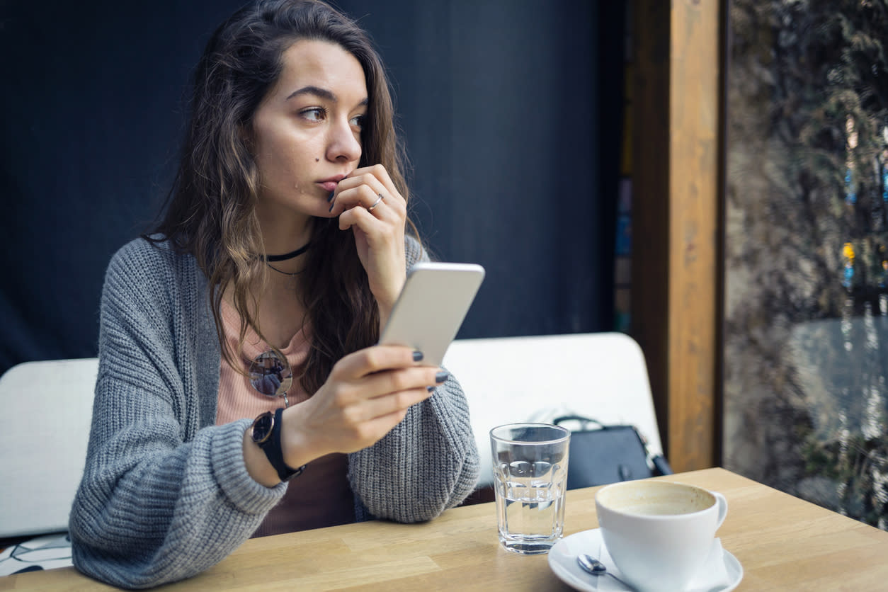 Woman sitting at a table at a coffee shop. She has her phone in her hand and is staring into the distance, looking anxious