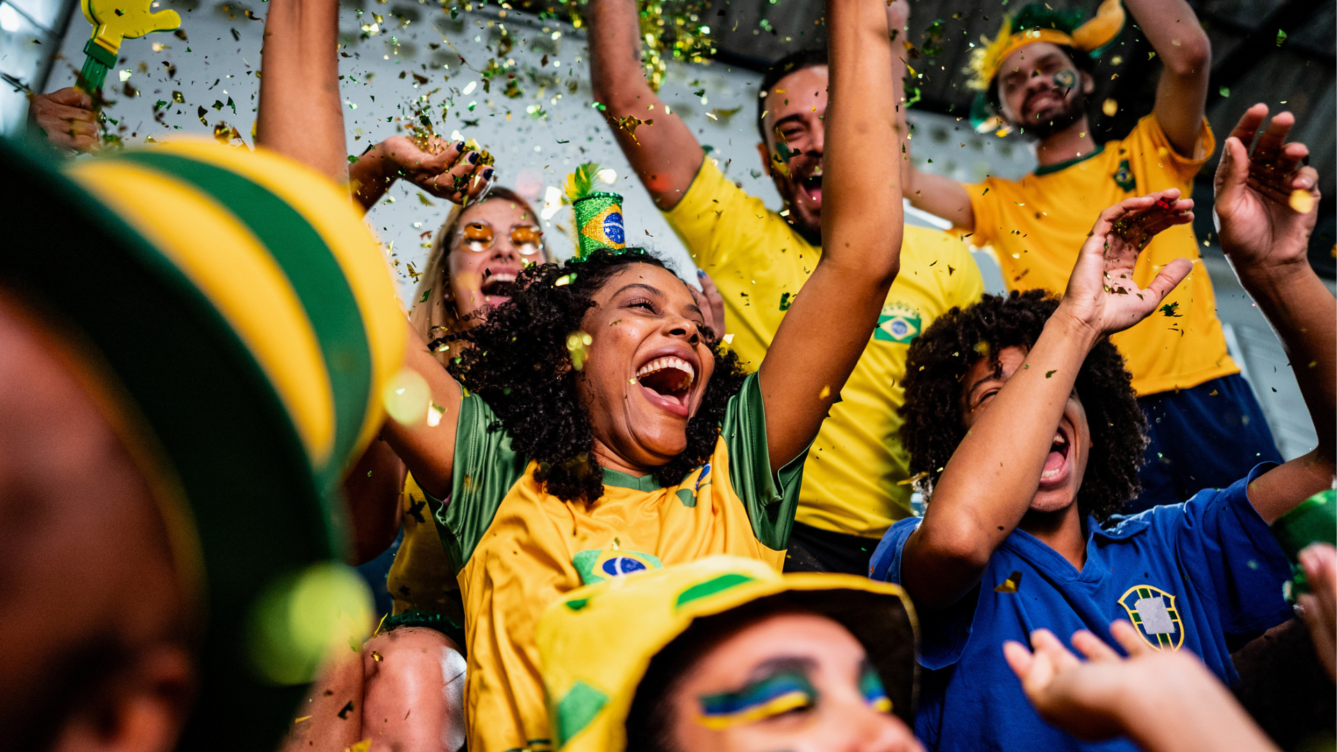 Crowd of Brazilian soccer fans wearing yellow and green jerseys celebrate enthusiastically, cheering with raised arms as confetti falls around them during a festive match-day gathering.