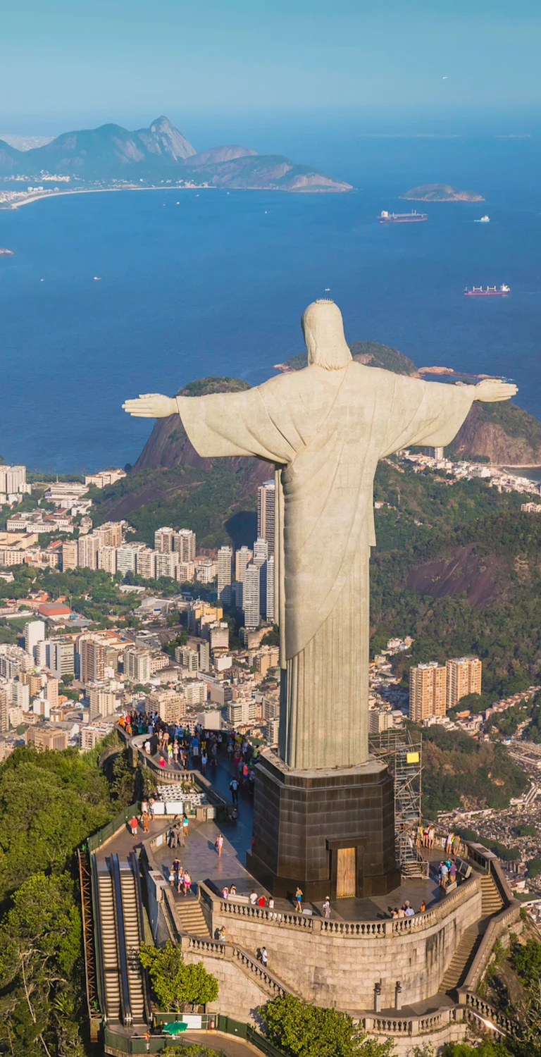 Aerial view of Rio de Janeiro, Brazil, featuring the Christ the Redeemer statue atop Corcovado Mountain overlooking the city, Guanabara Bay, Sugarloaf Mountain, and surrounding coastline under clear daylight.