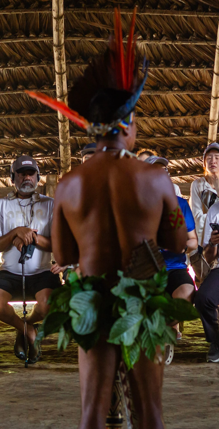 Visitors sit inside a large thatched-roof shelter, watching an Indigenous performer wearing a feathered headdress and leaf skirt. The performer stands with their back to the camera during a cultural demonstration.