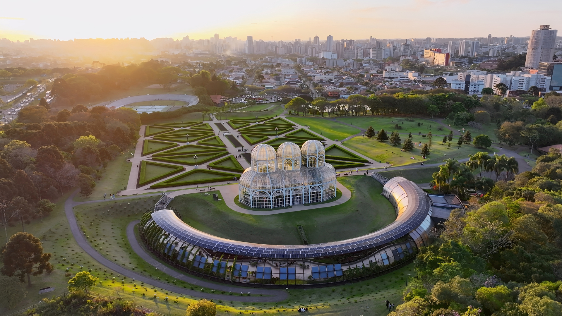 Aerial view of a glass greenhouse surrounded by formal geometric gardens and green lawns at sunset, with a large city skyline in the background and tree-lined parkland extending across the landscape.