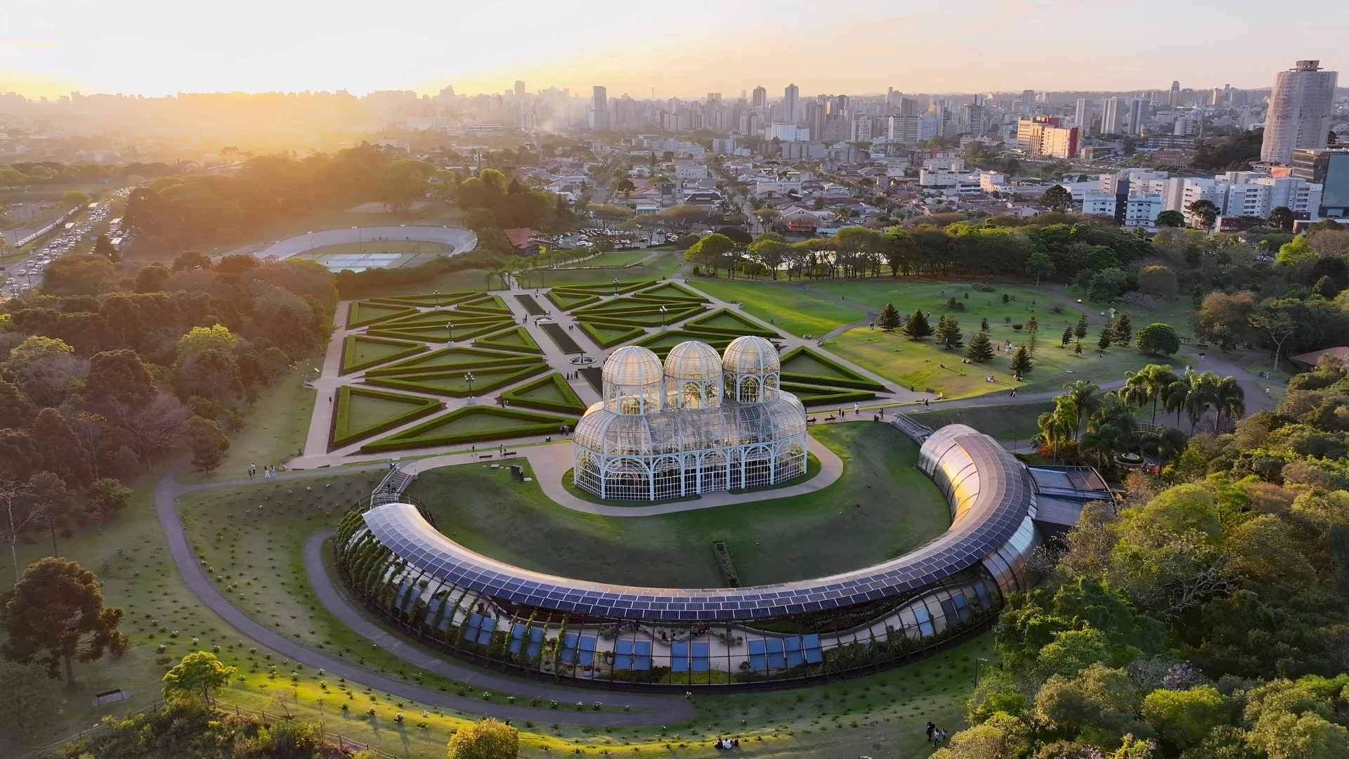 Aerial view of a glass greenhouse surrounded by formal geometric gardens and green lawns at sunset, with a large city skyline in the background and tree-lined parkland extending across the landscape.