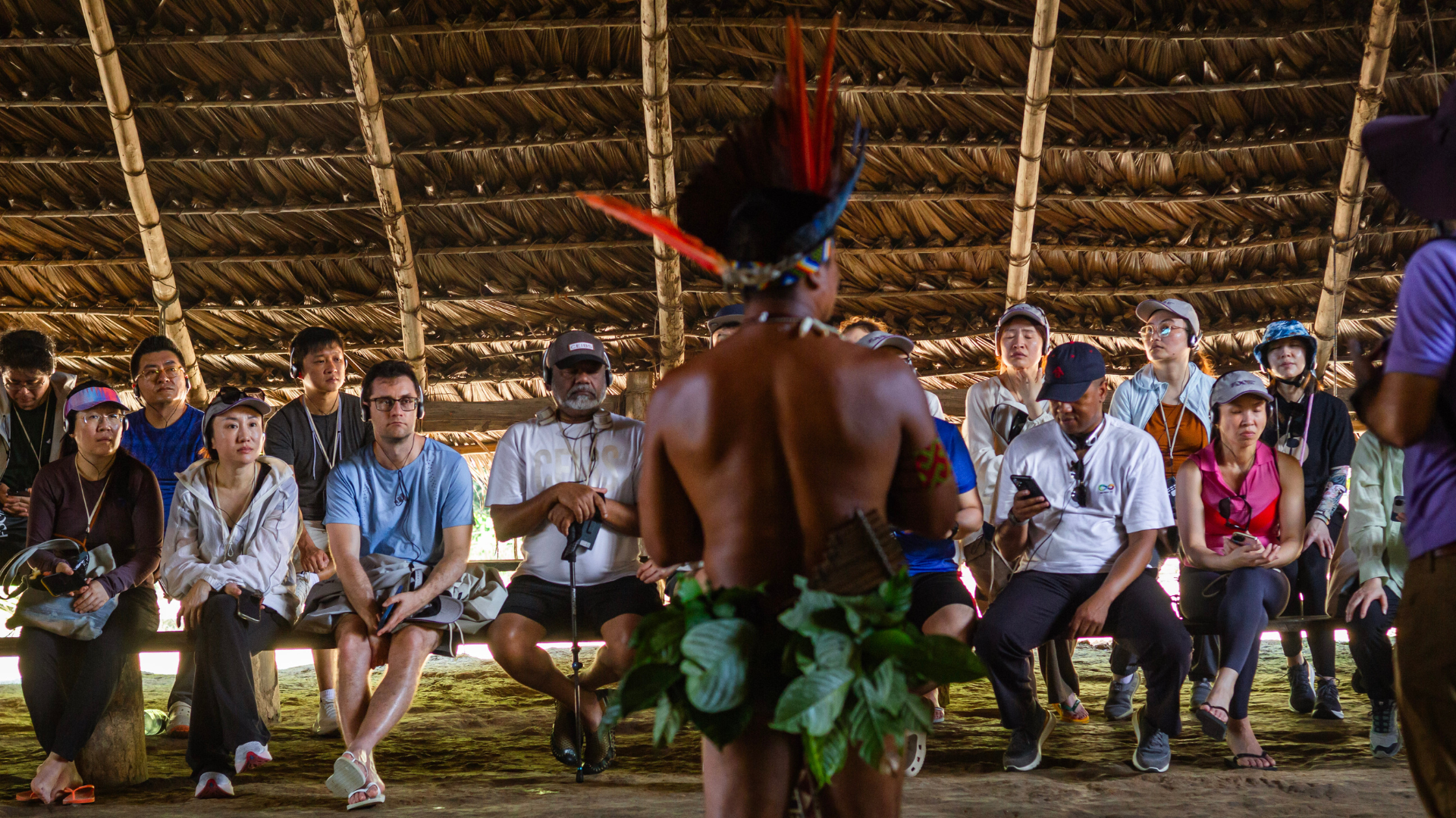 Visitors sit inside a large thatched-roof shelter, watching an Indigenous performer wearing a feathered headdress and leaf skirt. The performer stands with their back to the camera during a cultural demonstration.