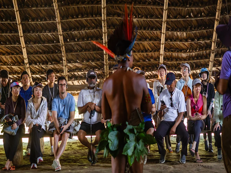 Visitors sit inside a large thatched-roof shelter, watching an Indigenous performer wearing a feathered headdress and leaf skirt. The performer stands with their back to the camera during a cultural demonstration.