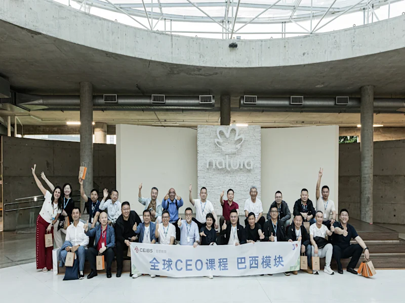 Group photo of participants at a global CEO program posing indoors in a modern building, smiling and waving behind a banner with Chinese text. The event stage and architectural concrete structure are visible in the background.