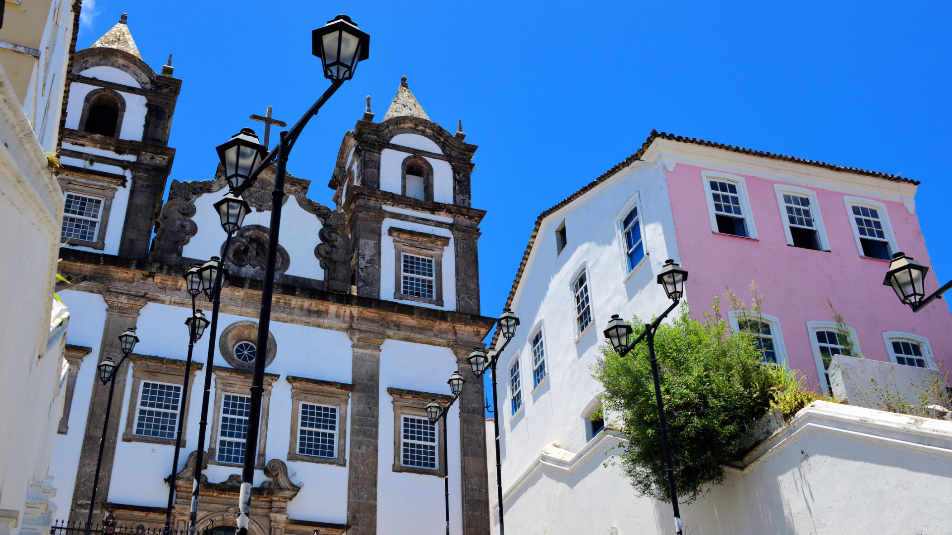 Historic church and colorful colonial buildings in Salvador, Brazil, viewed from a low angle. Black street lamps line the scene, while bright blue skies and white façades highlight the city's rich architectural heritage.