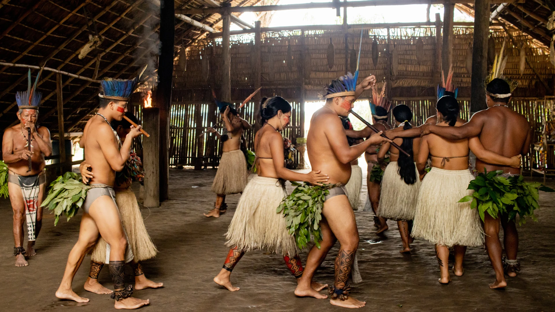 Indigenous community members perform a traditional dance inside a thatched communal house, wearing ceremonial attire and carrying leafy branches.