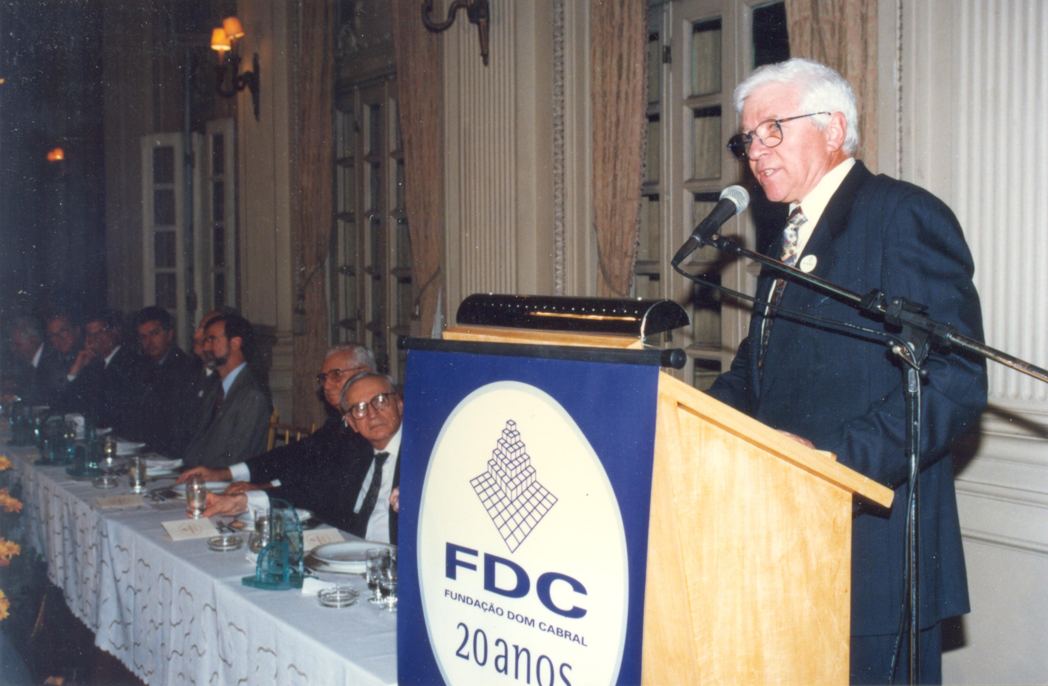Homem idoso falando em público na cerimônia de 20 anos da Fundação Dom Cabral, com uma mesa de convidados ao fundo em ambiente elegante.