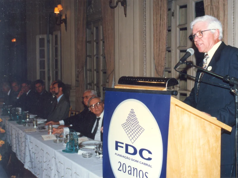 Homem idoso falando em público na cerimônia de 20 anos da Fundação Dom Cabral, com uma mesa de convidados ao fundo em ambiente elegante.