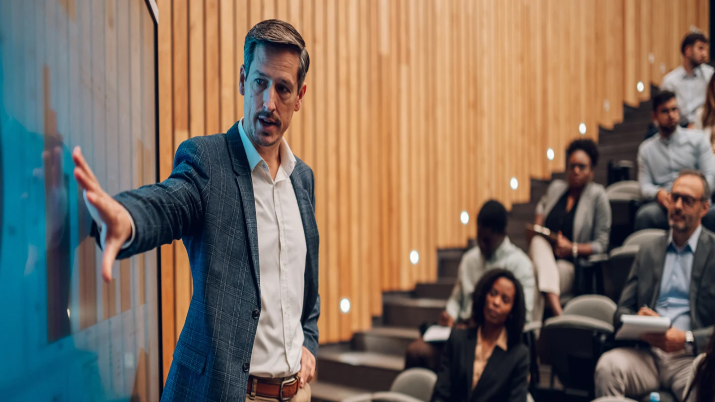 Homem de terno apresenta informações em uma tela interativa na frente de uma sala de aula ou auditório moderno, enquanto várias pessoas sentadas em arquibancadas acompanham a apresentação e fazem anotações.