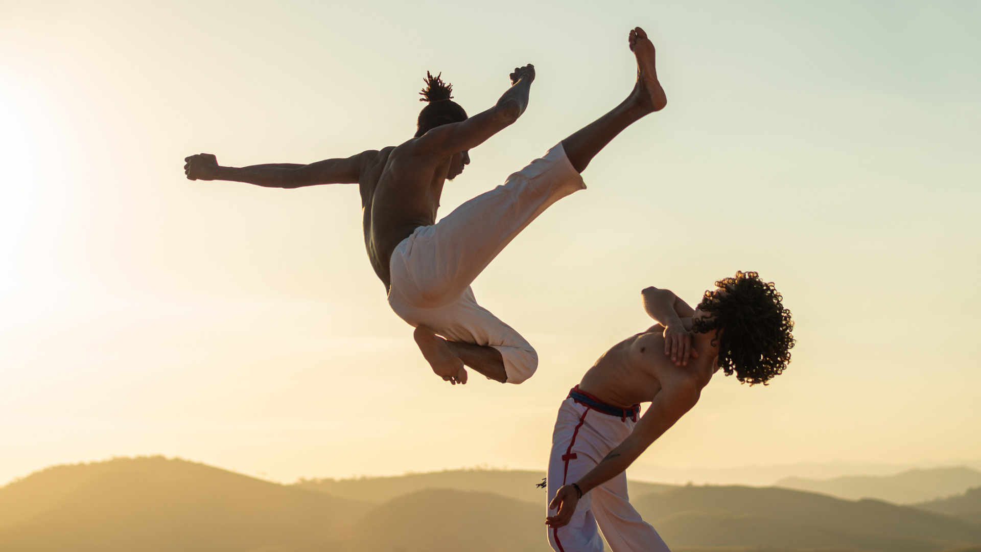 Silhouetted capoeira practitioners performing an acrobatic kick and dodge at sunset, with rolling hills in the background and warm golden light highlighting movement, balance, and Brazilian cultural tradition.