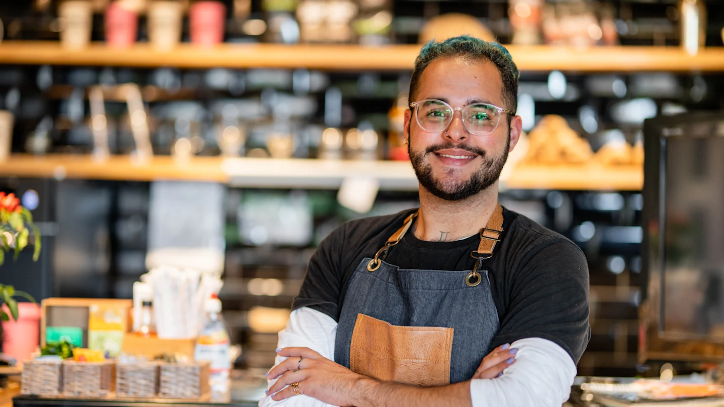 Homem de óculos e avental em frente a uma cafeteria, sorrindo e pronto para atender os clientes.