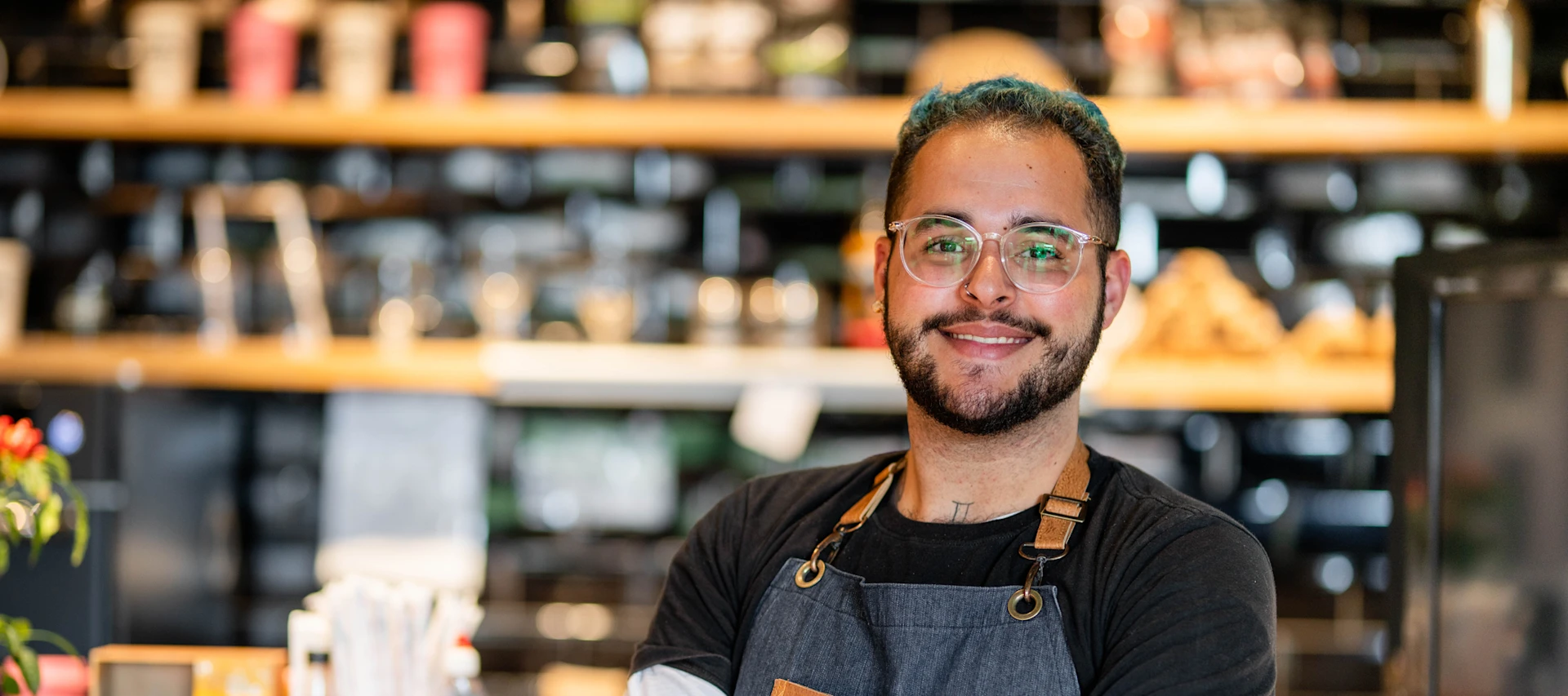 Homem de óculos e avental em frente a uma cafeteria, sorrindo e pronto para atender os clientes.