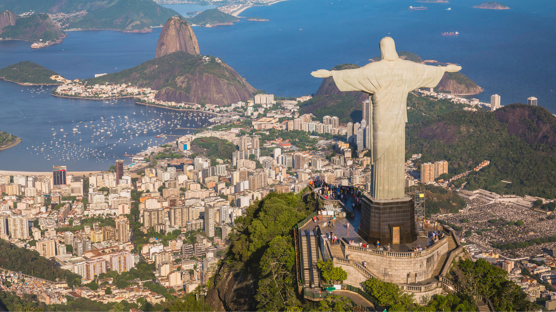 Aerial view of Rio de Janeiro, Brazil, featuring the Christ the Redeemer statue atop Corcovado Mountain overlooking the city, Guanabara Bay, Sugarloaf Mountain, and surrounding coastline under clear daylight.