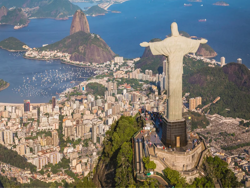 Aerial view of Rio de Janeiro, Brazil, featuring the Christ the Redeemer statue atop Corcovado Mountain overlooking the city, Guanabara Bay, Sugarloaf Mountain, and surrounding coastline under clear daylight.