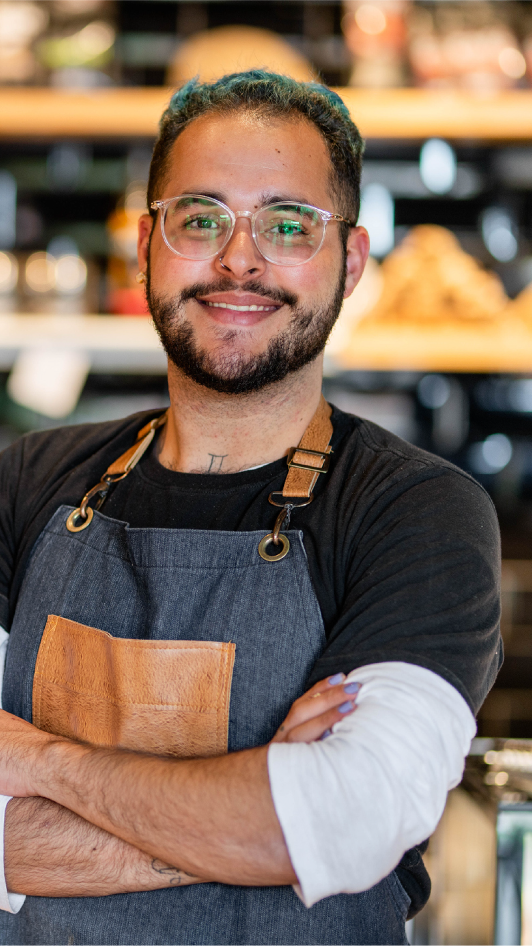 Homem de óculos e avental em frente a uma cafeteria, sorrindo e pronto para atender os clientes.