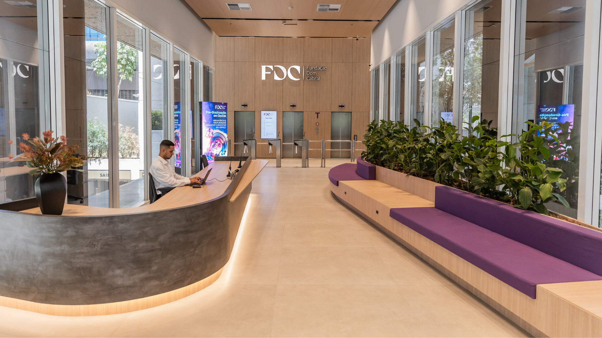 Modern office lobby with a curved reception desk, a staff member working at a computer, purple cushioned seating, indoor plants, floor-to-ceiling glass walls, and security turnstiles beneath an FDC-branded wood-paneled wall.