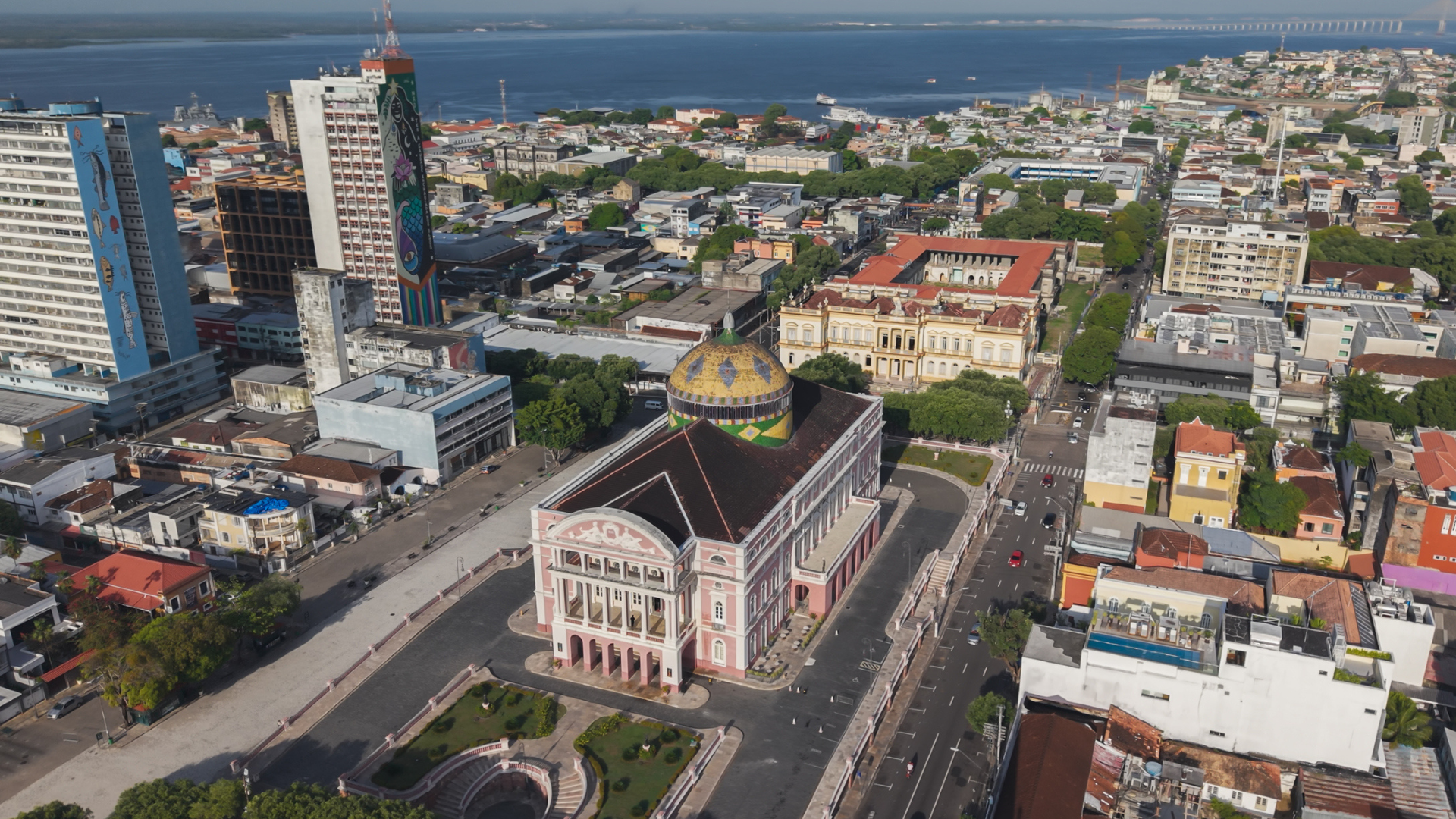 Aerial view of the historic Teatro Amazonas in downtown Manaus, featuring its colorful domed roof, surrounding city streets and buildings, with the Rio Negro visible in the background.